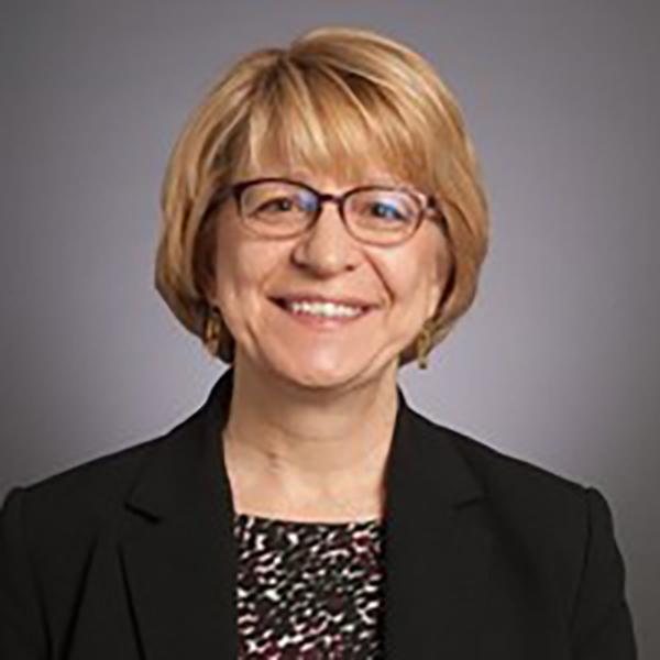 Professional image of a woman wearing a floral shirt, 黑色的外套, and black reading glasses, sitting against a grey background.